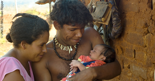 um homem, uma mulher e um bebê indígenas. O homem segura o bebê no colo.